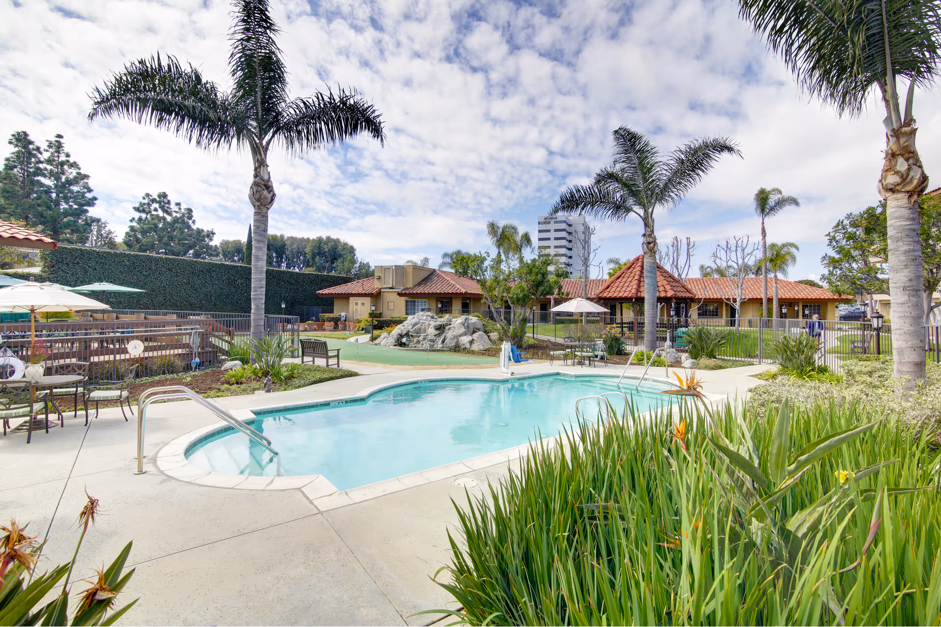 Outdoor area of Huntington Terrace featuring a swimming pool surrounded by palm trees, patio tables with umbrellas, benches, and a well-maintained garden. In the background, there are buildings with red-tiled roofs and a partly cloudy sky.