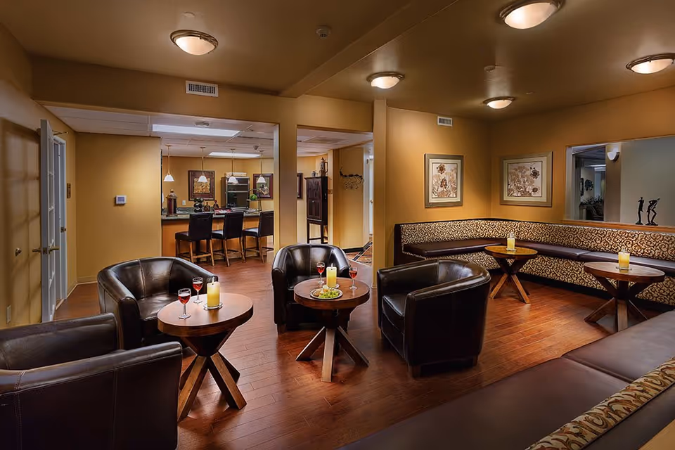 Cozy communal lounge with leather armchairs, round wooden tables holding drinks and candles, banquette seating along the wall, and a bar area visible in the background.