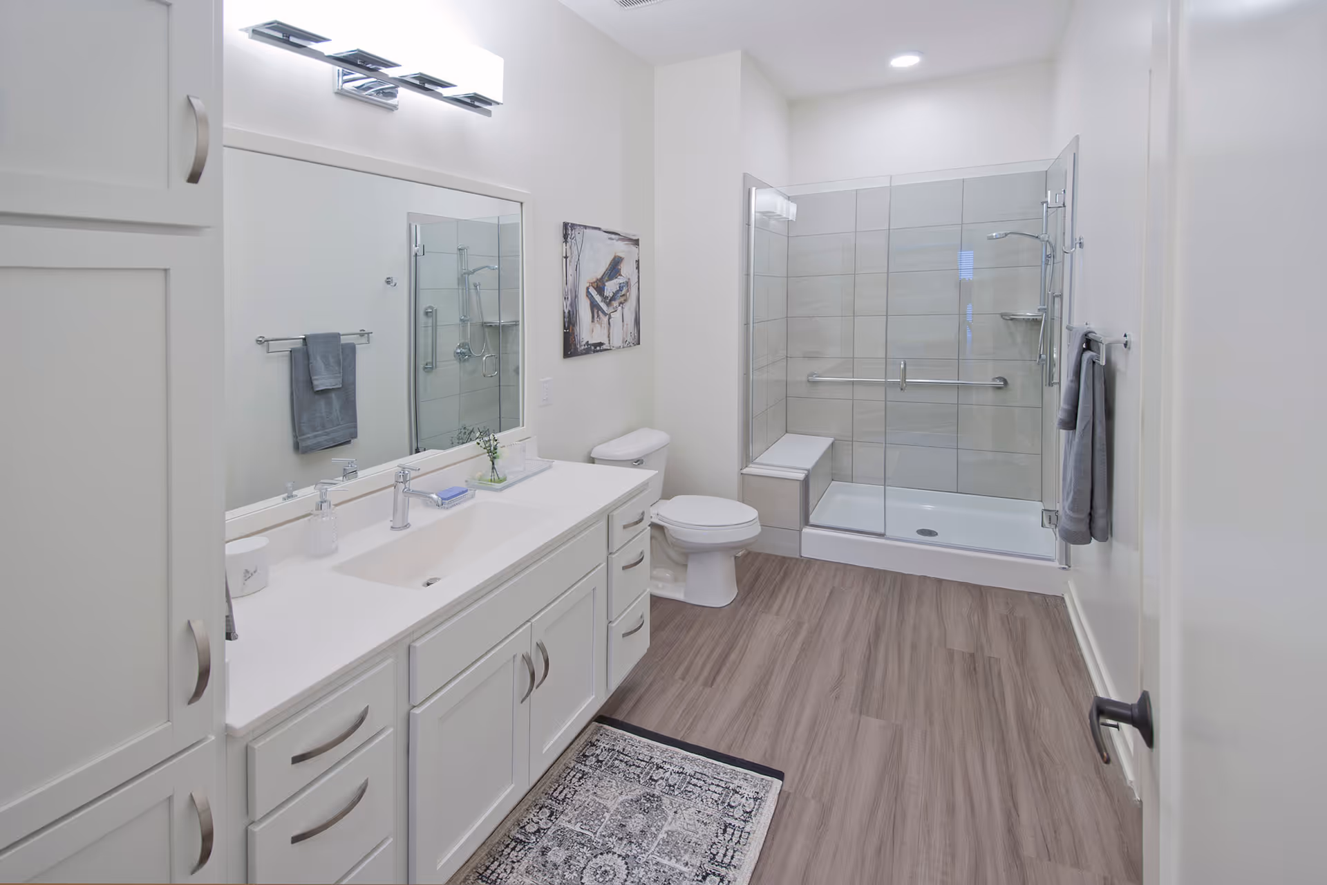 A modern bathroom with a large vanity featuring a white countertop and multiple drawers, a wide mirror with a light fixture above it, a toilet, and a glass-enclosed shower with a built-in bench and grab bars. The floor has wood-like tiles and there are gray towels hanging on the walls.