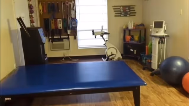 A rehabilitation room with a blue therapy table in the center, exercise equipment including a stationary bike, therapy bands hanging on the wall, a large exercise ball, and medical devices on stands. The room has wooden flooring and a window with a white blind.