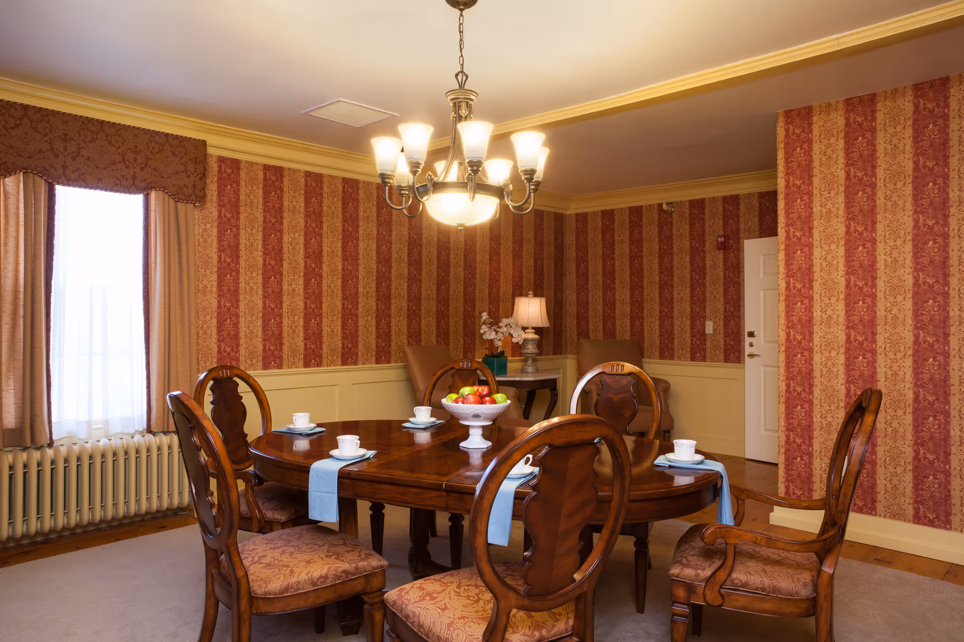Formal dining room with a round wooden table set with teacups and a fruit bowl, upholstered chairs, chandelier, and red patterned wallpaper.