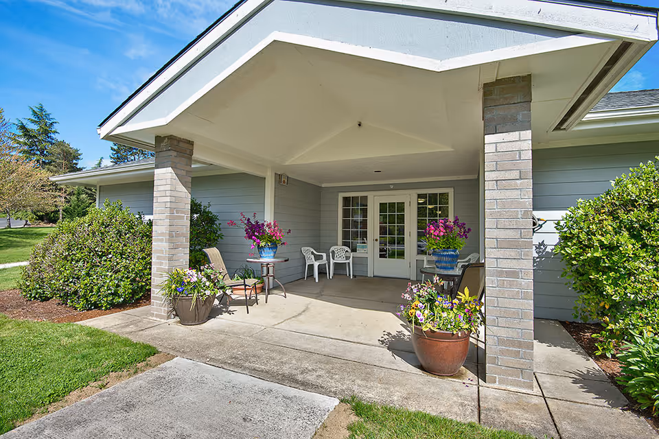 Covered entrance area of a building with two brick pillars, potted flowers, chairs, and a glass double door leading inside. The surrounding area has green bushes and a well-maintained lawn under a clear blue sky.