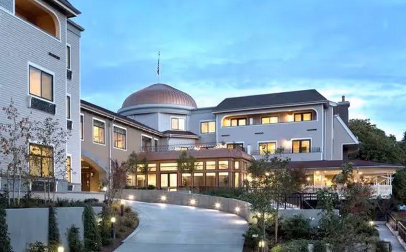 Exterior view of a multi-story senior living facility building with a curved driveway leading up to the entrance. The building features a dome-shaped roof section, multiple windows, and outdoor landscaping with trees and shrubs under a partly cloudy sky.