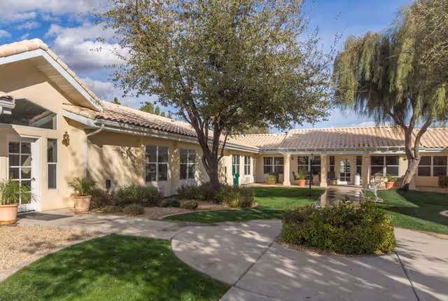 Outdoor courtyard area of a senior living facility with beige stucco buildings, tiled roofs, green grass, trees, shrubs, and a paved walkway under a partly cloudy blue sky.