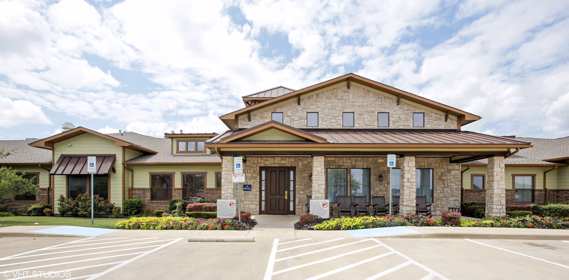 Front exterior of an assisted living and memory care building with a stone facade, covered entrance with rocking chairs, landscaped beds, and accessible parking spaces.