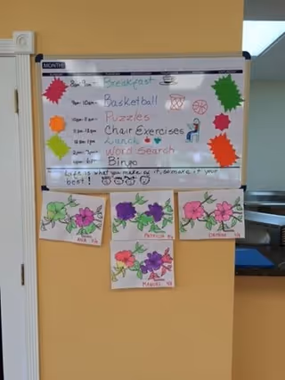 A whiteboard mounted on a yellow wall displays a daily activity schedule including breakfast, basketball, puzzles, chair exercises, lunch, word search, and bingo. Below the schedule, there are four colorful drawings of flowers labeled with names. The room appears to be part of an indoor facility with a doorway visible on the left and a window or opening on the right.