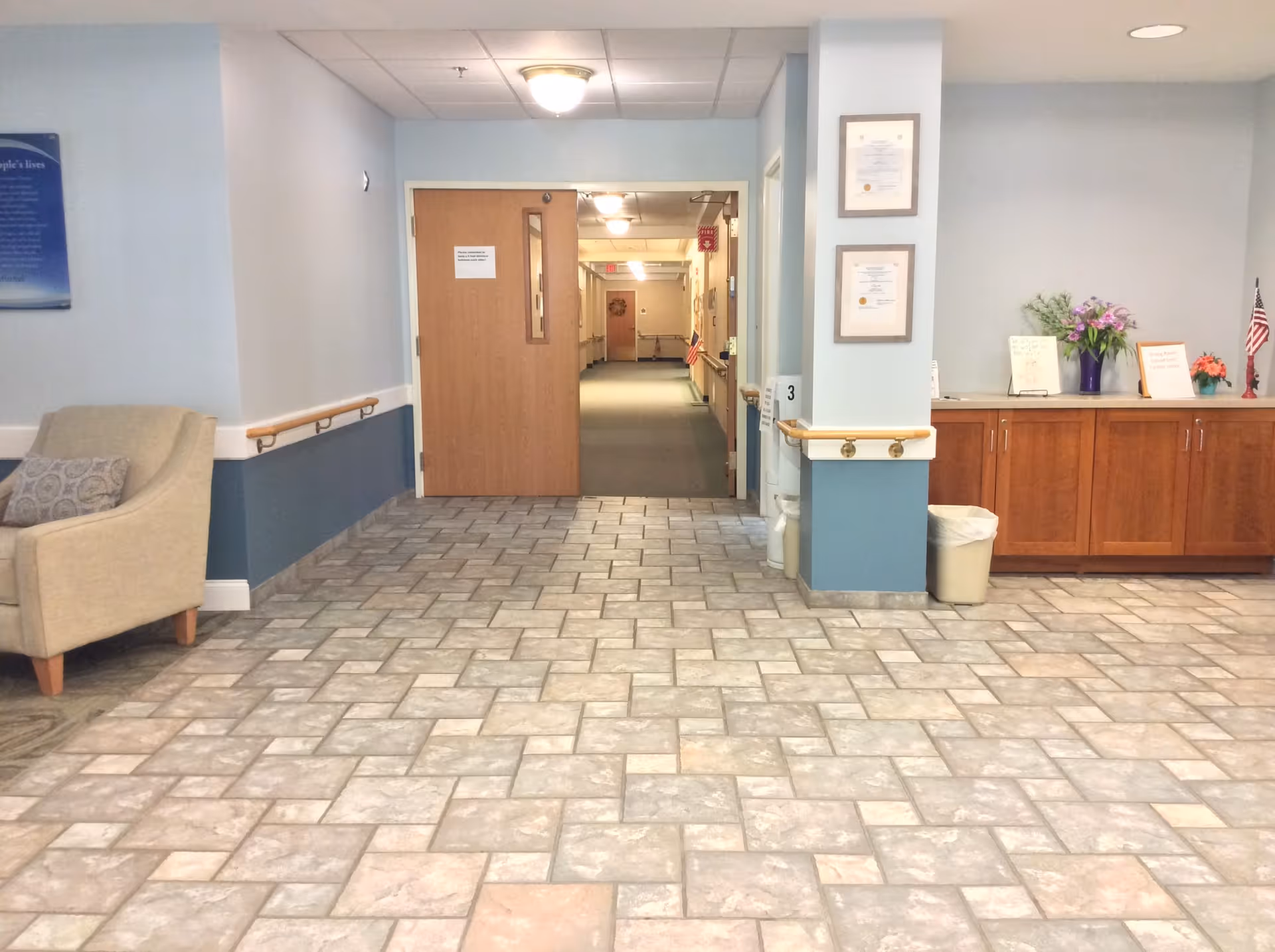 Interior view of a hallway in an assisted living facility with tiled floor, light blue walls, a beige armchair on the left, wooden cabinets with flowers and small American flags on the right, and a wooden door leading to a carpeted corridor.