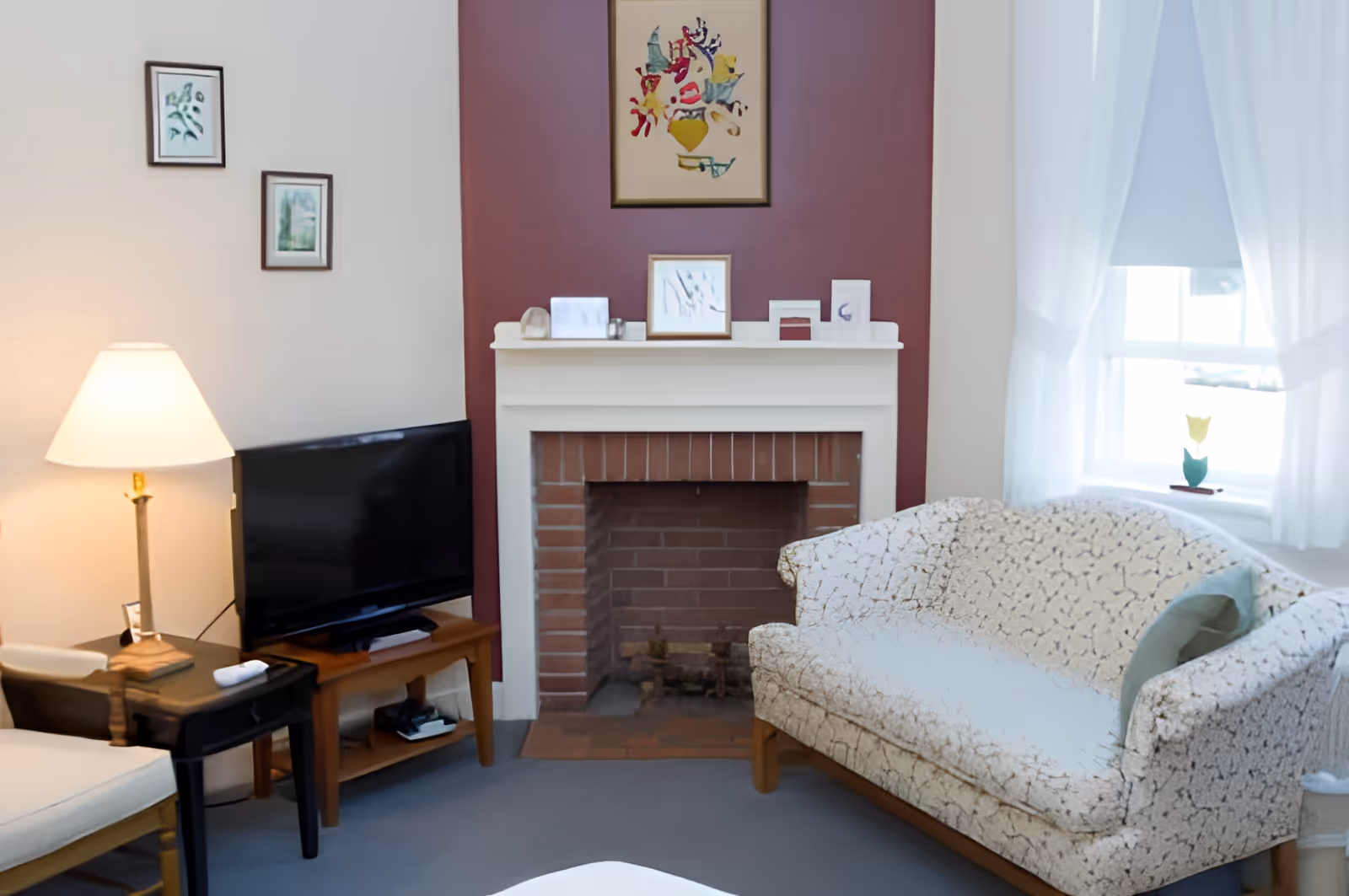 A cozy living room with a patterned loveseat, a brick fireplace with a white mantel, a flat-screen TV on a wooden stand, a side table with a lamp, and framed artwork on the walls. A window with sheer white curtains lets in natural light.
