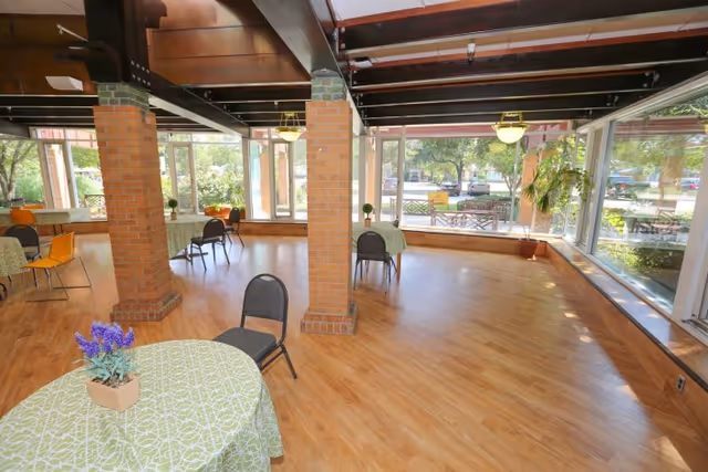 A spacious dining area with wooden floors, brick pillars, and large windows letting in natural light. Several tables covered with green patterned tablecloths and chairs are arranged around the room. There are potted plants on the tables and near the windows, with a view of trees and outdoor seating visible outside.