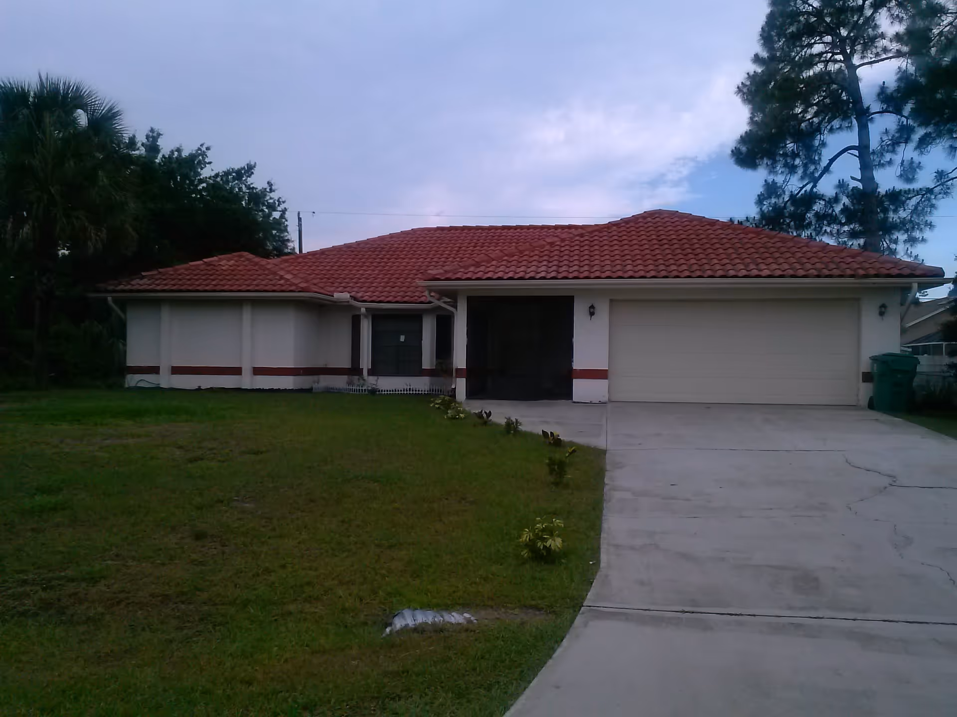Single-story house with a red tile roof, white exterior walls with a red stripe, a two-car garage, and a concrete driveway. The front yard has green grass and small plants lining the driveway. Trees are visible in the background under a cloudy sky.