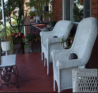 A porch area with white wicker chairs and a small table with a sewing machine base. There are potted plants and flowers along the railing, and a brick wall with windows in the background.