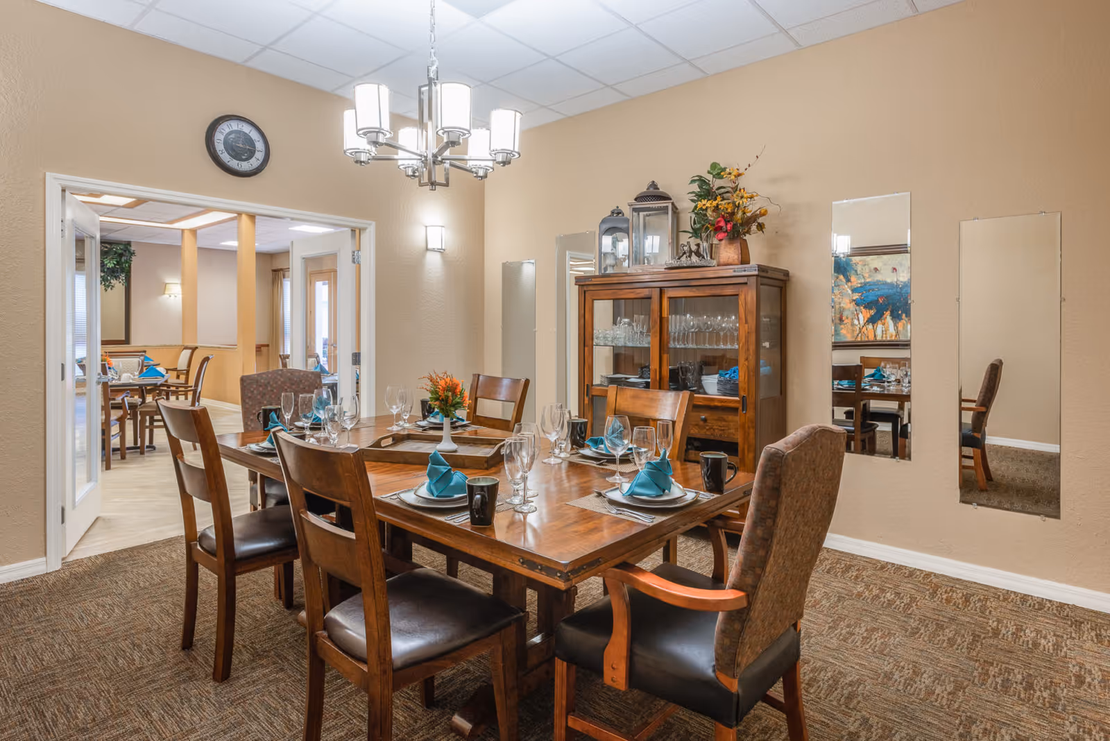 A dining room in a senior living facility with a wooden dining table set for six people. The table has plates, glasses, mugs, and teal-colored napkins. There is a wooden cabinet with glassware and decorative items against the wall. The room has beige walls, a carpeted floor, a chandelier, and three rectangular mirrors on the wall. An adjoining room with more dining tables and chairs is visible through an open doorway.