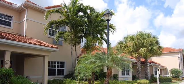 Exterior view of Sonata Delray Beach facility showing a beige building with red tile roof, multiple windows, and surrounded by palm trees and greenery under a partly cloudy sky.