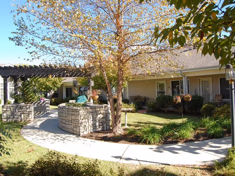 Outdoor courtyard area at a senior living facility with a curved concrete walkway, stone planters, a pergola, a tree with autumn leaves, and a single-story building with beige siding and white trim in the background.