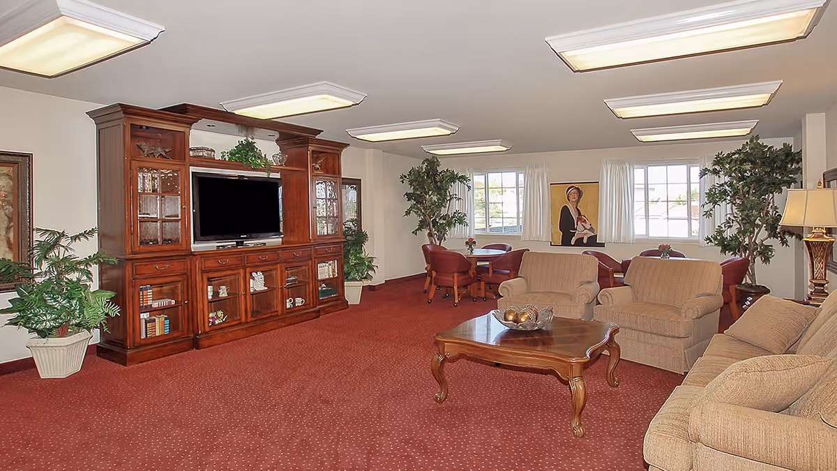 Communal lounge with sofas and armchairs facing a large wooden entertainment center and TV, a coffee table, dining table, and potted plants.