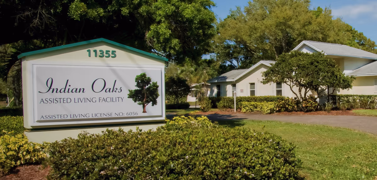 A roadside sign reading "Indian Oaks Assisted Living Facility" with low white buildings and landscaped shrubs and trees in the background.