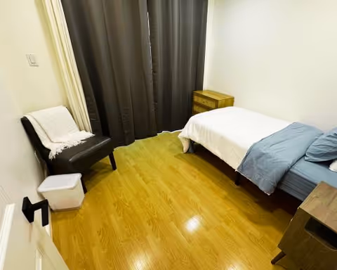 A small bedroom with a single bed covered with white and blue bedding, a wooden nightstand, a dark chair with a white throw blanket, a small white trash bin, and dark curtains covering the window. The floor is wooden and the walls are white.