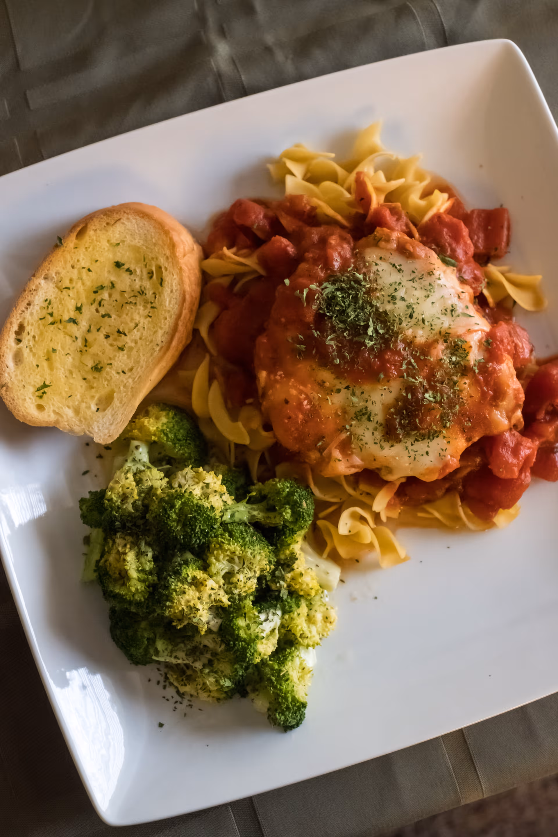 A white square plate with a serving of cooked broccoli, a slice of garlic bread, and a portion of egg noodles topped with tomato sauce and melted cheese garnished with herbs.