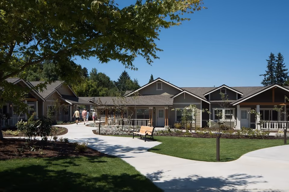 Outdoor view of a senior living facility with a paved walkway, green lawn, trees, and shrubs. Several single-story buildings with porches are visible under a clear blue sky. Three people are walking along the path.