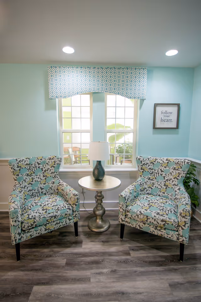 A cozy seating area with two floral patterned armchairs in shades of blue, white, and gray, positioned on either side of a small round table with a decorative lamp. Behind the chairs is a window with a blue and white patterned valance, and a framed wall art that reads 'follow your heart.' The walls are painted light blue and the floor has a wood-like finish.