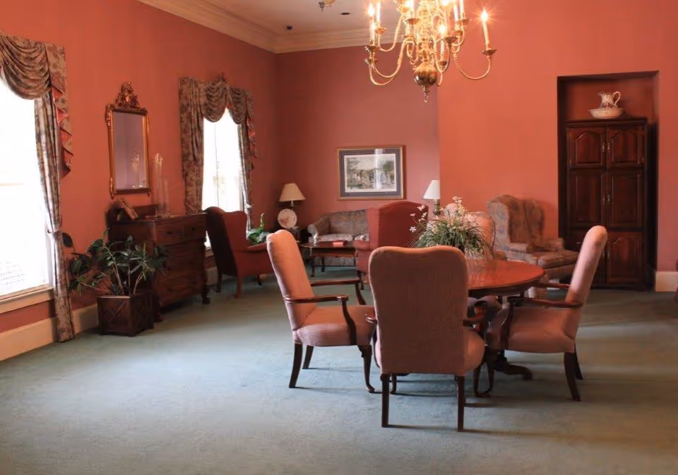 A cozy living room with pink walls and green carpet featuring a round wooden table with four upholstered chairs in the center. There are two large windows with floral curtains on the left, a wooden chest of drawers with a mirror above it, and several armchairs and a sofa arranged around a coffee table. A chandelier hangs from the ceiling, and a wooden cabinet with decorative items is against the far wall.