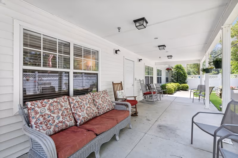 Covered outdoor patio area with white siding walls, multiple windows, and a concrete floor. The patio is furnished with a cushioned wicker sofa with patterned pillows, several rocking chairs, and metal chairs. There are hanging plants and greenery visible beyond the patio, along with a white fence enclosing the area.