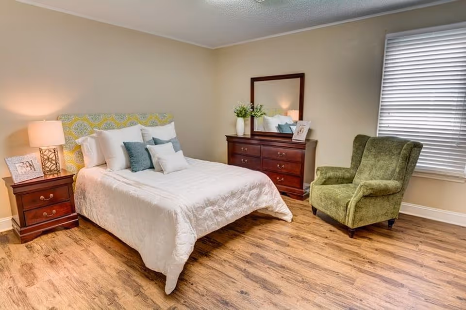 A cozy bedroom with a bed featuring a patterned yellow and green headboard, white bedding, and several pillows. Next to the bed is a wooden nightstand with a lamp and a framed photo. Across from the bed is a wooden dresser with a mirror, decorative vases, and a framed picture. A green upholstered armchair is positioned near a window with closed blinds. The room has wood flooring and beige walls.