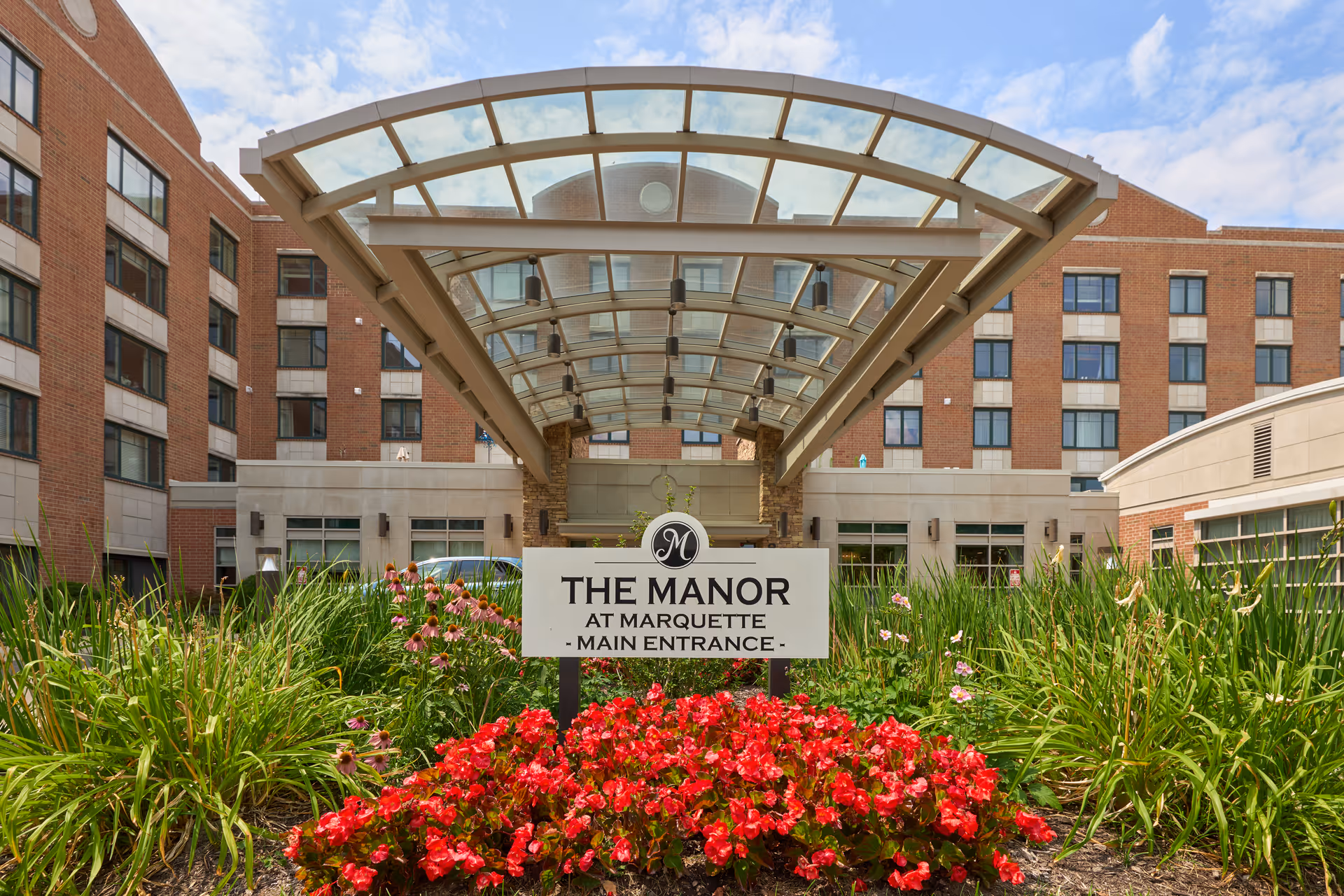 The main entrance of The Manor at Marquette, featuring a covered glass canopy above the entrance driveway. The building is a multi-story brick structure with numerous windows. In front of the entrance, there is a garden with red and pink flowers and green plants surrounding a sign that reads 'THE MANOR AT MARQUETTE - MAIN ENTRANCE -'.