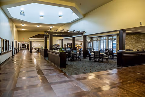 Spacious interior of a senior living facility with a wide hallway featuring wooden flooring and a large skylight above. To the right, there is a dining area with multiple tables and chairs, separated by dark wooden beams and partial walls. The space is well-lit with natural light coming through large windows and ceiling lights.