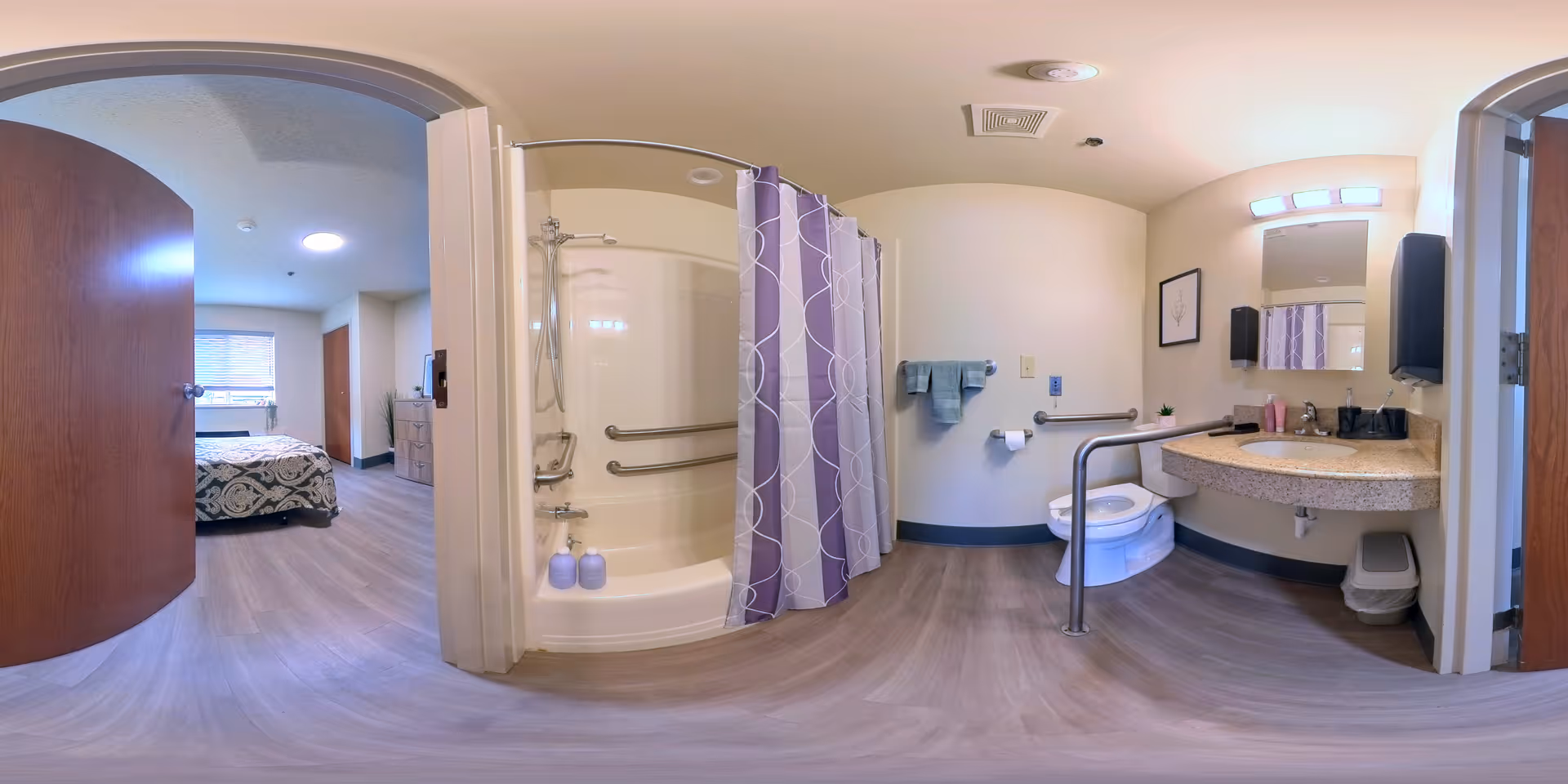 A wide-angle view of an accessible bathroom connected to a bedroom. The bathroom features a shower with grab bars and a purple patterned shower curtain, a toilet with support rails, a countertop with a sink, a mirror, and hand towels. The bedroom visible through the open door has a bed with patterned bedding and a window with blinds.