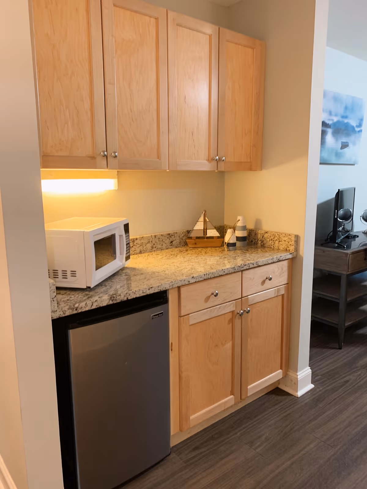 A small kitchenette area with light wood cabinets, a granite countertop, a white microwave, and a small stainless steel refrigerator. Decorative items including a small wooden sailboat and two striped vases are placed on the countertop. In the background, part of a living room with a TV and a painting on the wall is visible.