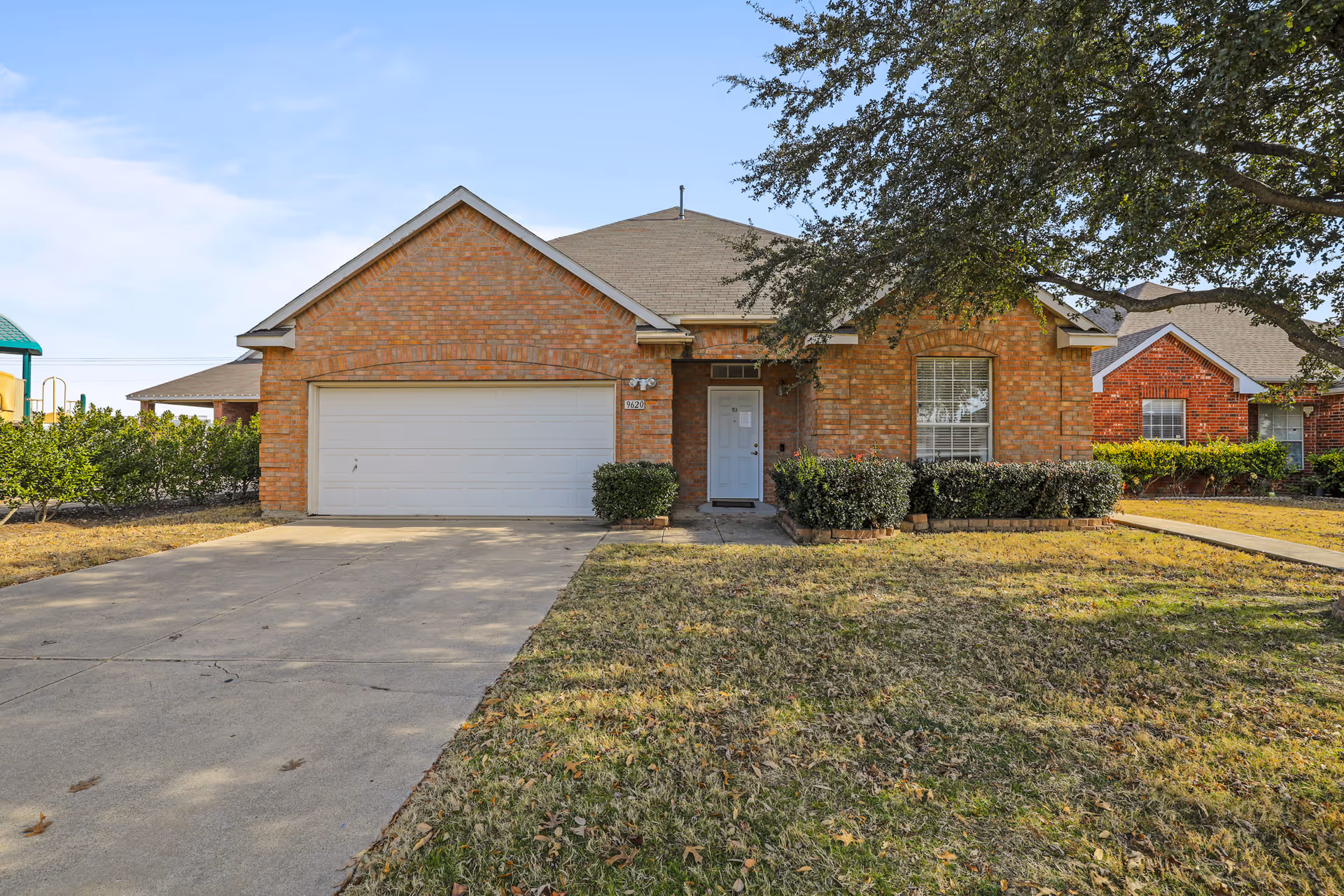 Front exterior view of a single-story brick house with a two-car garage, a driveway, and a lawn with some bushes and a tree.