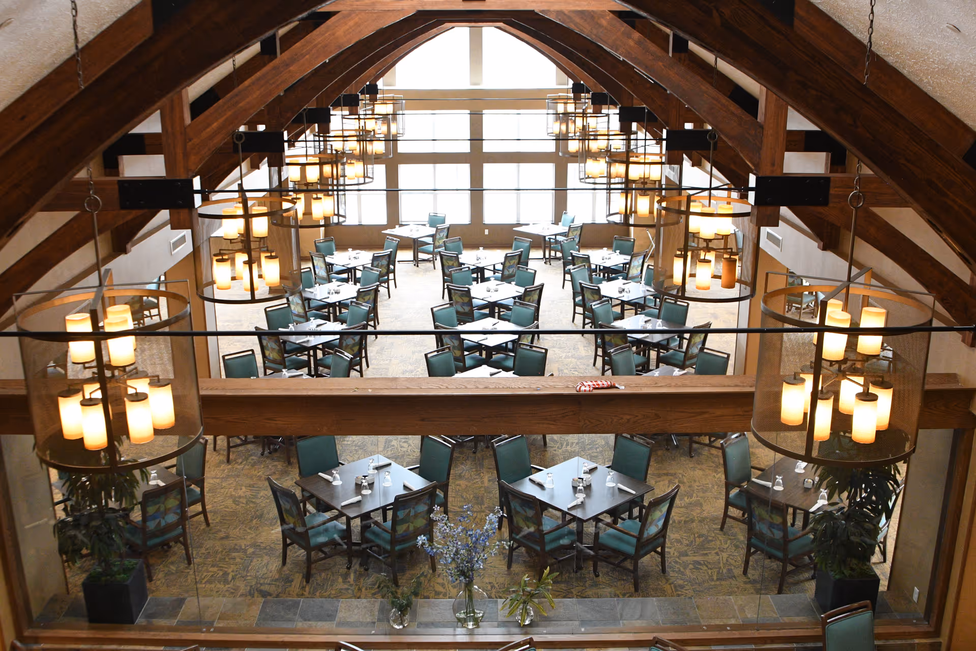 Spacious dining room with multiple tables and green chairs beneath wooden vaulted beams and hanging chandeliers.
