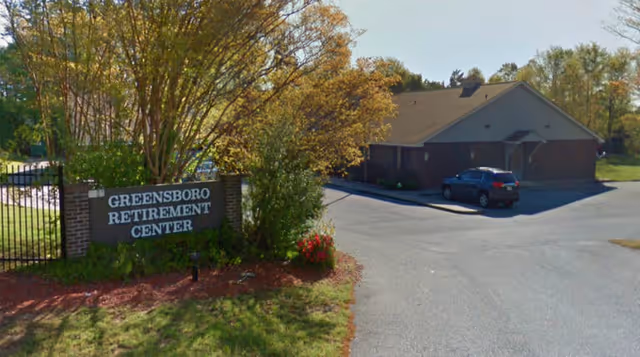 Exterior view of Greensboro Retirement Center showing a brick sign with the facility name surrounded by trees and bushes, with a building and a parked car visible in the background under a clear sky.