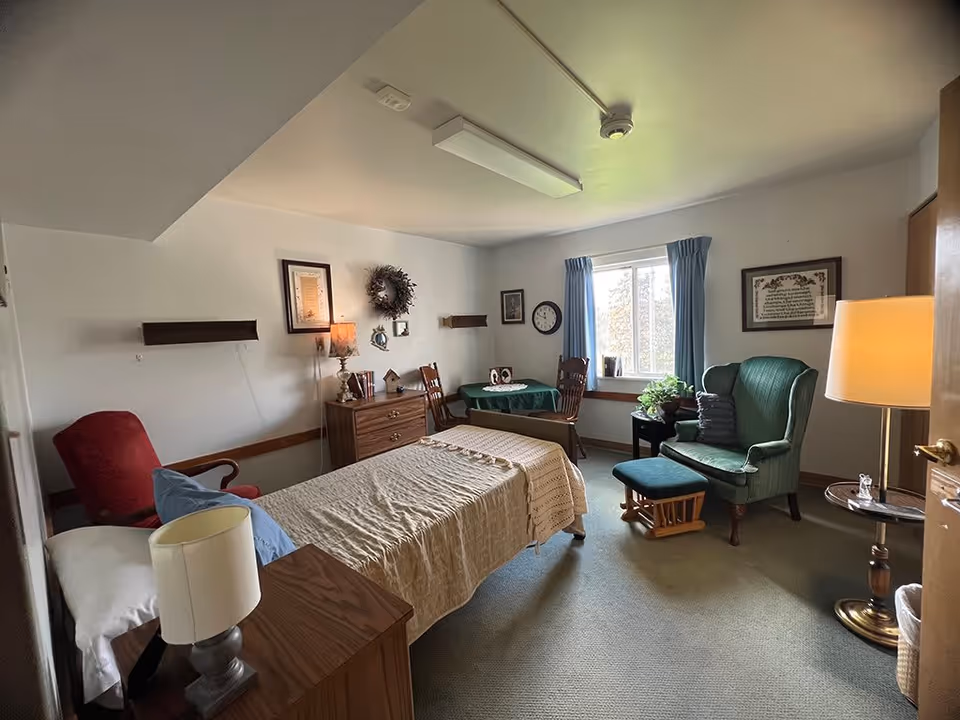 A cozy bedroom in a senior living facility with a single bed covered with a beige blanket, a red armchair, a green wingback chair with a matching footrest, a small round table with two wooden chairs, a wooden dresser with a lamp and decorative items, framed pictures and a wreath on the walls, a window with blue curtains, and a standing lamp near the door.