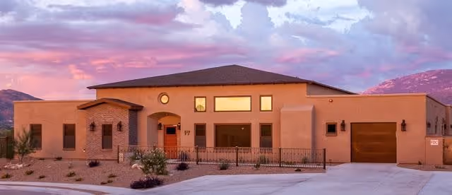 Exterior view of a single-story building with a southwestern architectural style, featuring a flat roof, beige stucco walls, and a small fenced front patio. The sky is colorful with pink and purple hues during sunset, and mountains are visible in the background.