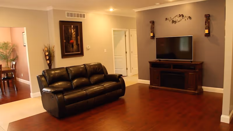 A living room with a black leather sofa facing a flat-screen TV mounted on a wooden TV stand against a gray accent wall. The room has wooden flooring and beige walls with white trim. There is a decorative wall piece above the TV and two candle holders on either side. To the left, there is an open doorway leading to a dining area with a wooden dining table and chairs, and a plant in the corner.