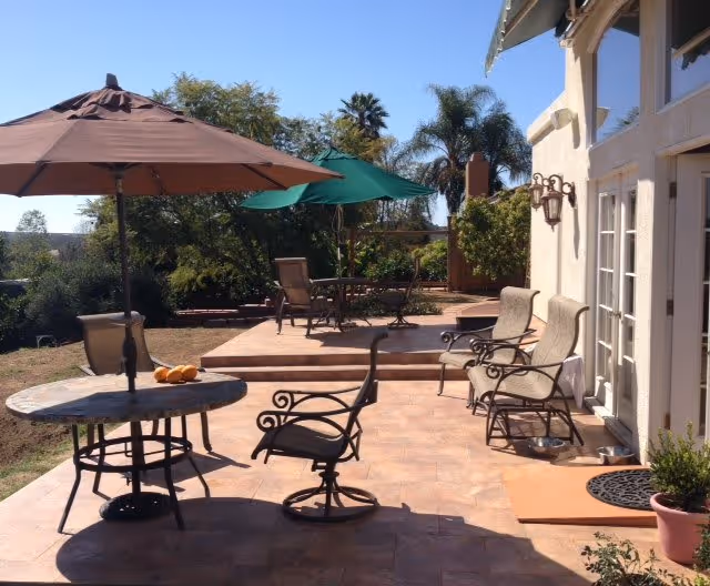 Outdoor patio area with several metal chairs and tables, two large umbrellas providing shade, potted plants, and a view of trees and clear sky in the background.
