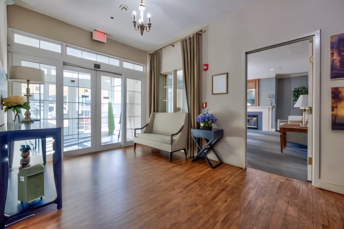 A bright interior room with wooden flooring, a beige loveseat against the wall, a small black side table with a vase of purple flowers, and large glass doors leading to an outdoor area. A chandelier hangs from the ceiling, and through an open doorway, another room with a fireplace and furniture is visible.