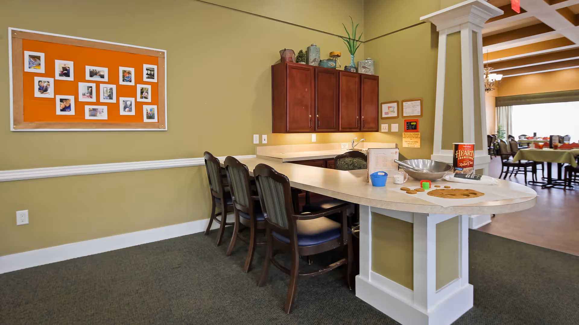 Interior view of a communal area in Meadowood Assisted Living and Memory Care featuring a curved countertop with four chairs, a bulletin board with photos on the wall, wooden cabinets, and a glimpse into a dining area with tables and chairs in the background.