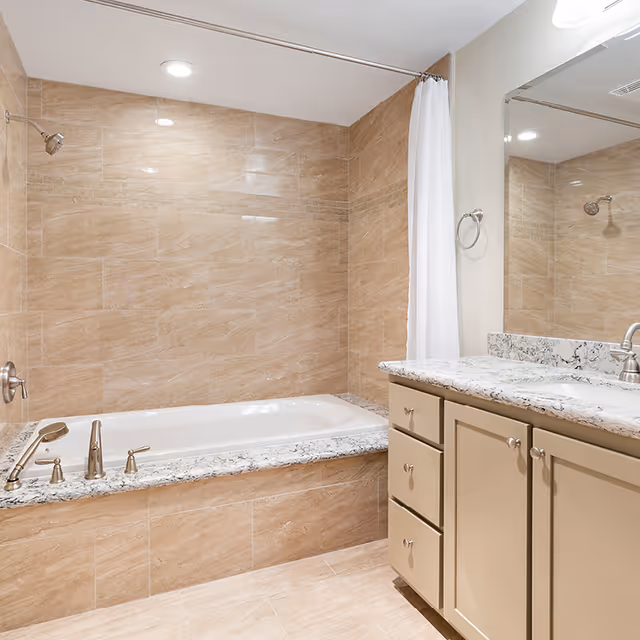 A modern bathroom featuring a built-in bathtub with beige tiled walls and floor. The bathtub has a granite surround and is equipped with silver fixtures including a faucet and handheld showerhead. To the right, there is a vanity with a granite countertop, beige cabinets, and a large mirror above it. A white shower curtain hangs from a rod above the bathtub.