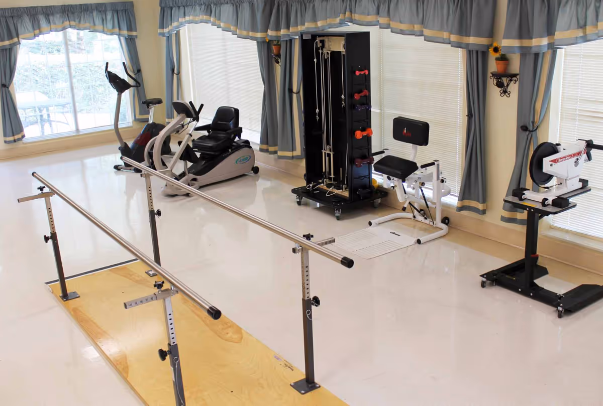 A rehabilitation exercise room with parallel bars on a wooden platform, a recumbent exercise bike, a multi-function weight machine, a seated exercise machine, and a hand cycle machine. The room has large windows with blue and beige curtains and a small potted sunflower on a wall shelf.