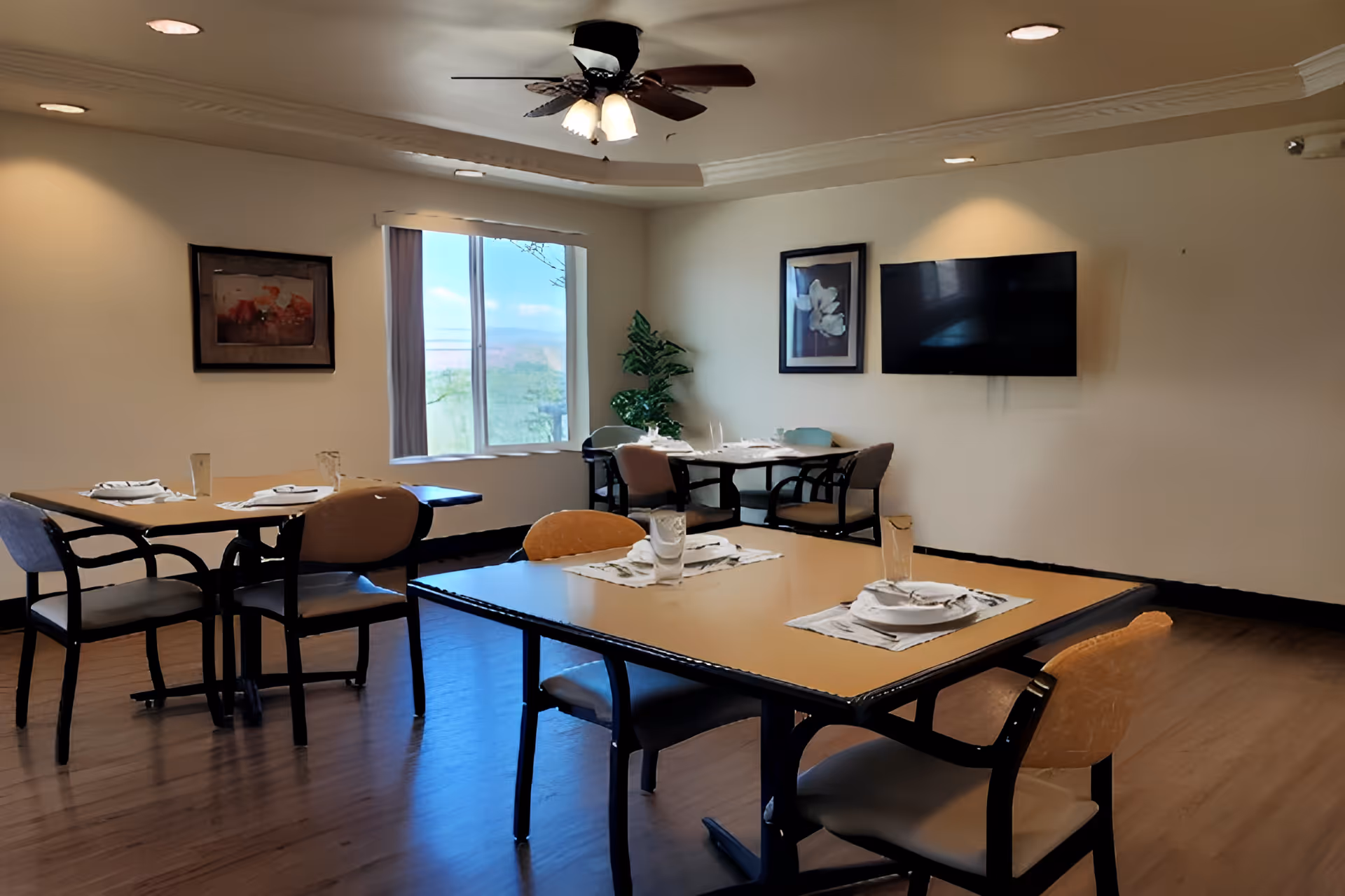 A dining room with several tables set with plates, glasses, and napkins. The room has wooden flooring, a ceiling fan with lights, framed artwork on the walls, a large window with a view outside, and a flat-screen TV mounted on the wall.