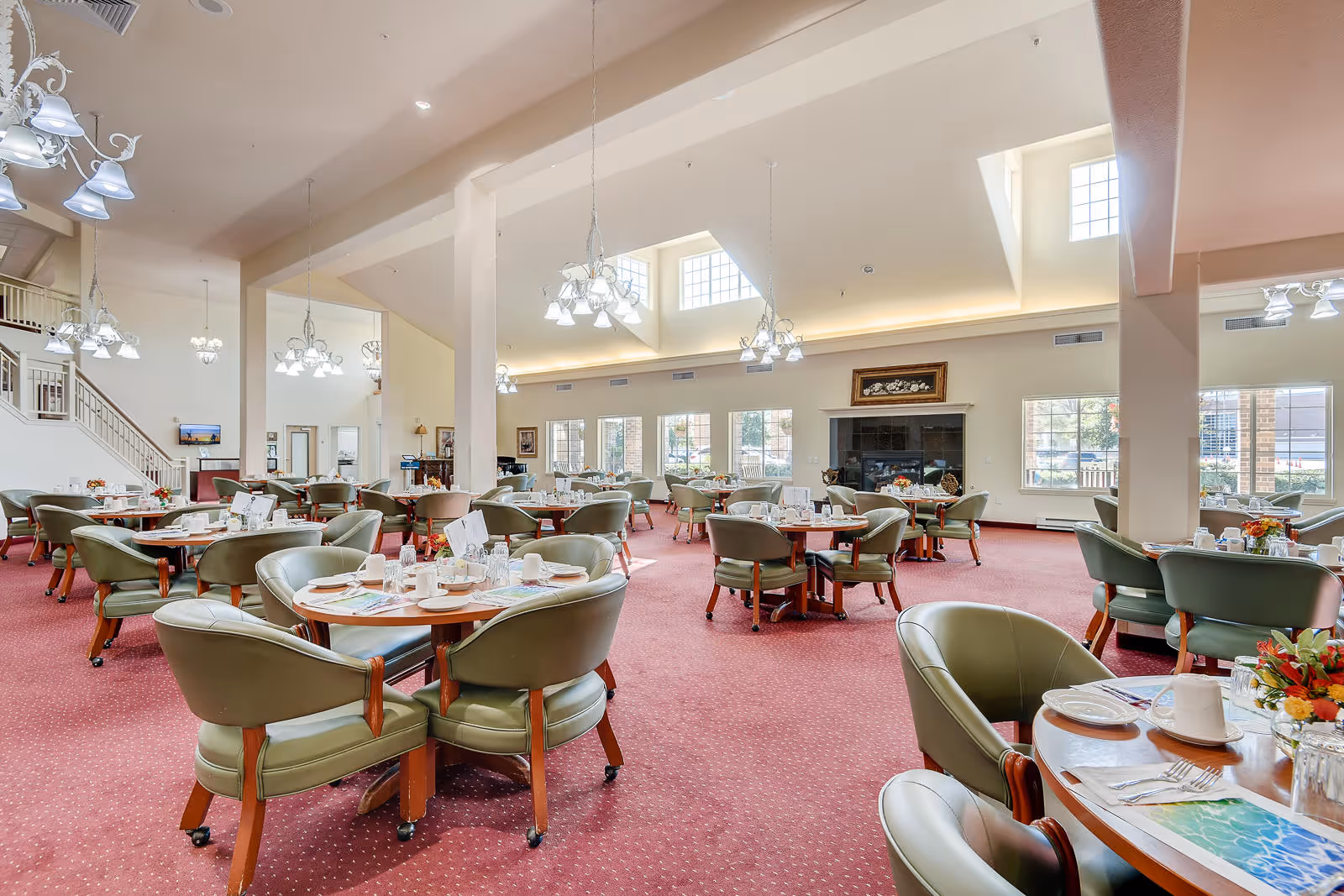 A spacious dining room with multiple round tables set for meals, surrounded by green cushioned chairs on a red carpet. The room features large windows allowing natural light, high ceilings with chandeliers, and a fireplace with a framed picture above it.