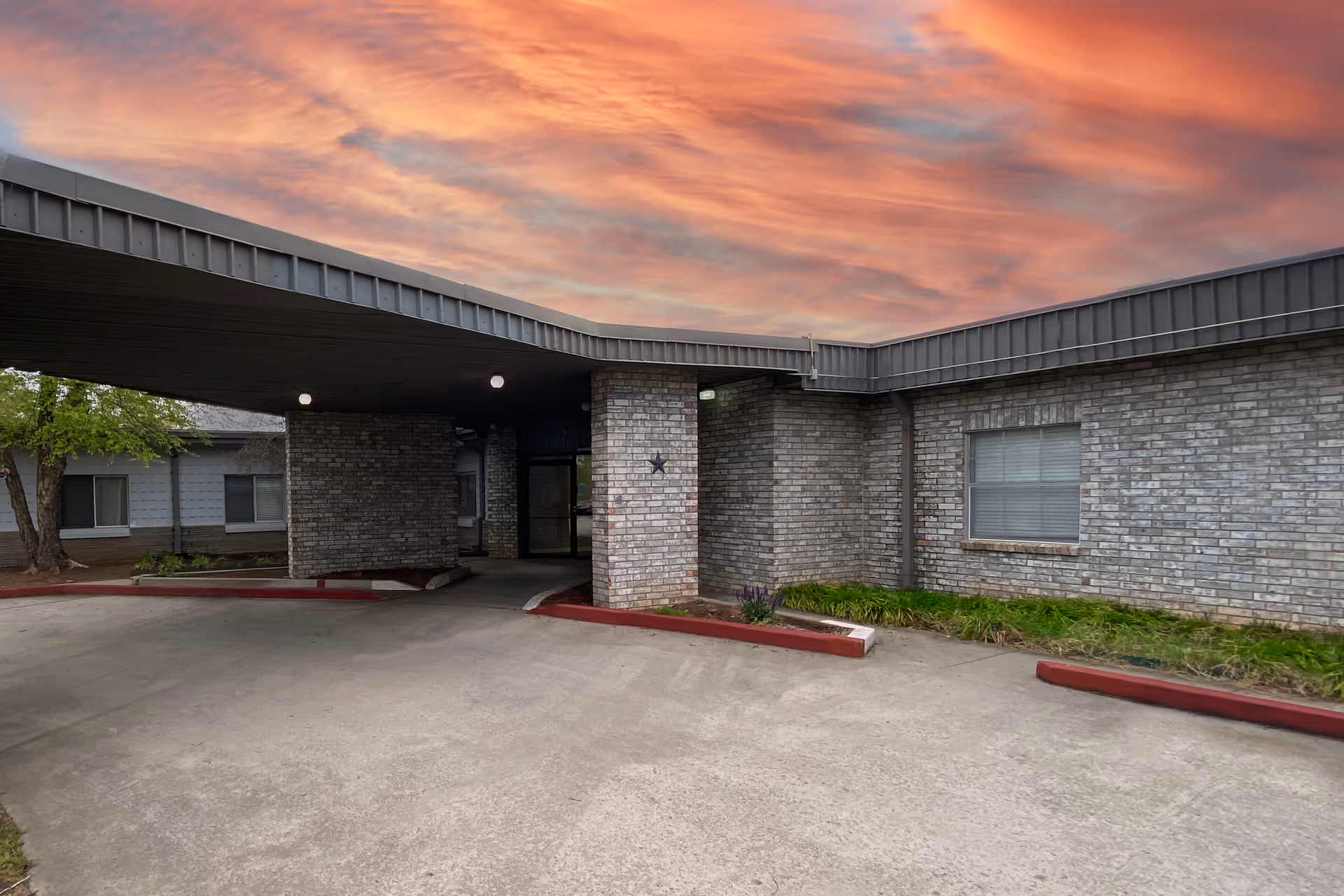 Exterior view of Oak Hills Care Center Skilled Nursing & Rehabilitation showing a covered entrance with brick walls and a driveway under a colorful sunset sky.