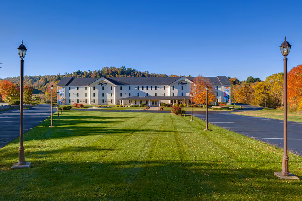 Front view of a large multi-story senior living building with a wide manicured lawn, lamp posts, and parking areas under a clear blue sky.
