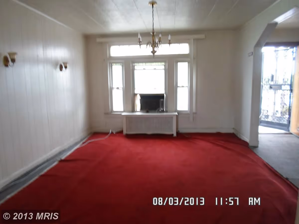 Empty room with red carpet, white walls, a window air conditioning unit in the middle window, two wall sconces on the left wall, a chandelier hanging from the ceiling, and an open doorway on the right leading to another area with a metal gate visible outside.