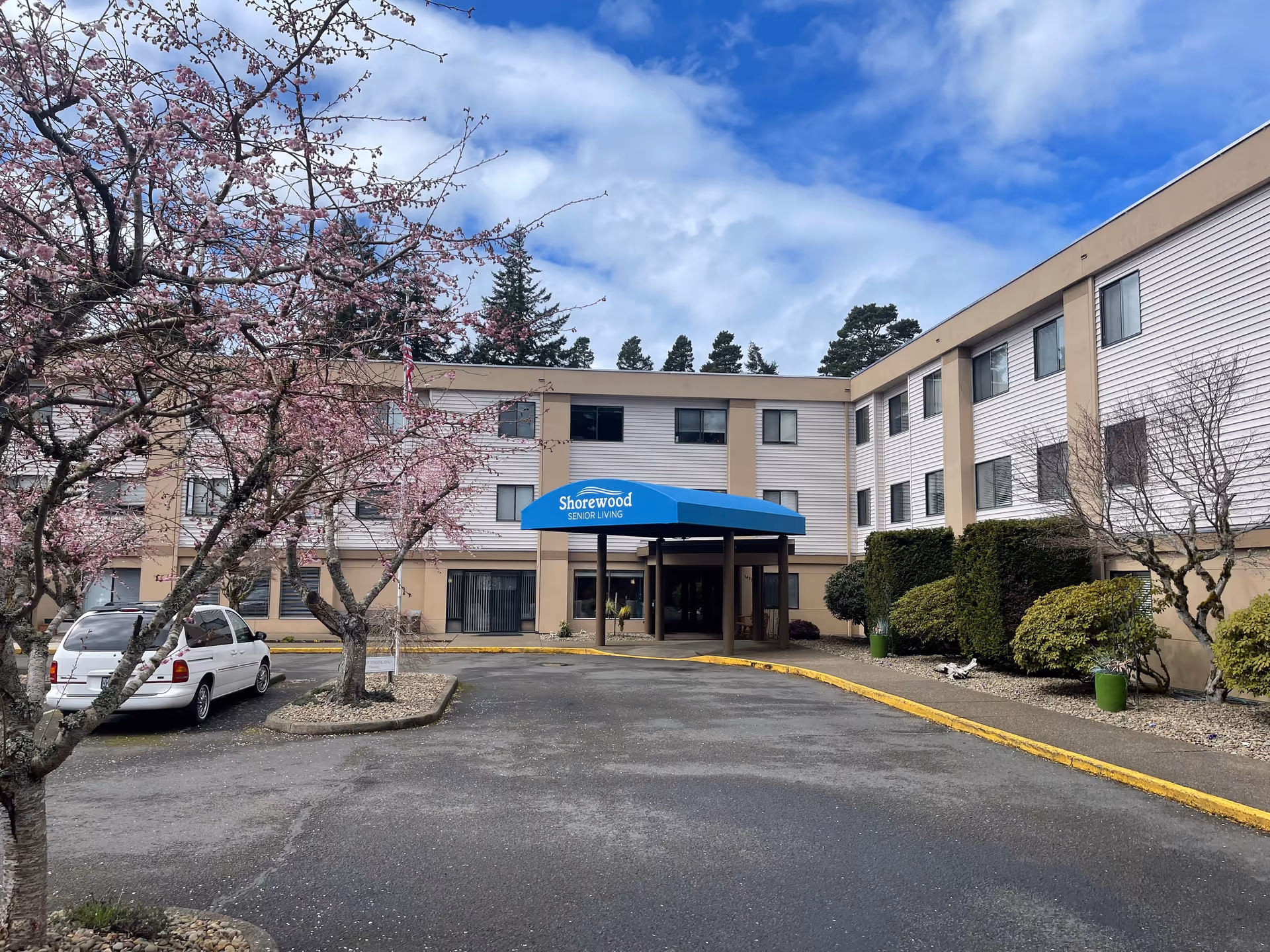 Exterior view of Shorewood Senior Living facility with a three-story building, a blue entrance canopy displaying the facility name, a driveway, a white parked car, and blooming pink trees under a partly cloudy sky.
