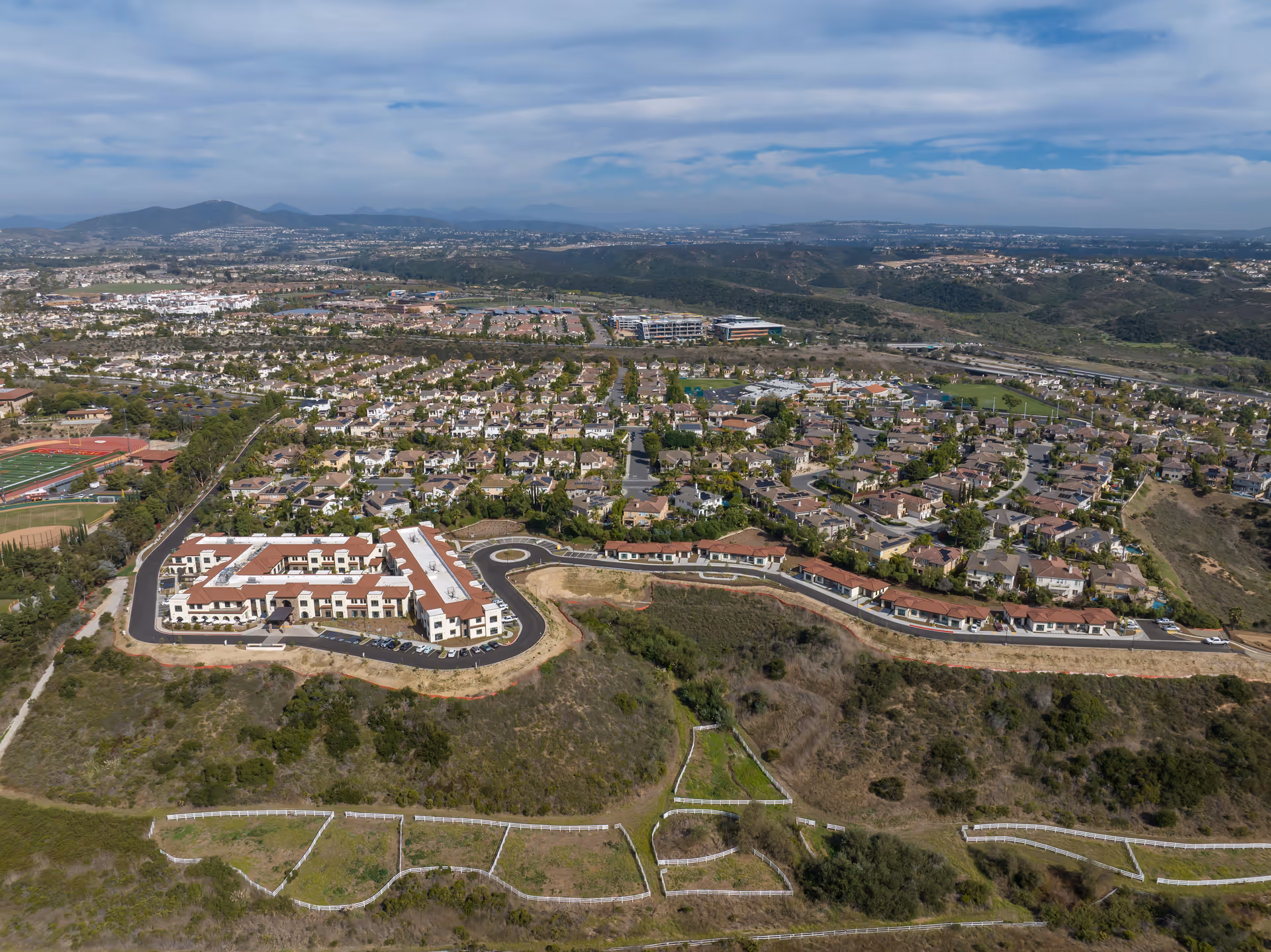 Aerial view of a residential area with a senior living facility named Westmont of Carmel Valley in the foreground, surrounded by hills and greenery, with a larger suburban neighborhood and mountains in the background under a partly cloudy sky.