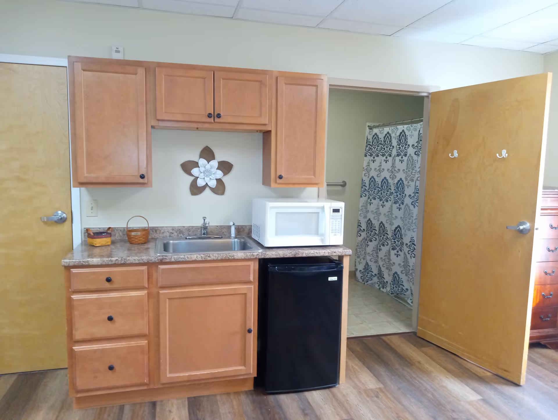 A small kitchenette area with wooden cabinets, a countertop with a sink, a white microwave, and a black mini refrigerator. To the right, an open door reveals a bathroom with a shower curtain featuring a blue and white pattern. The floor is wood laminate and the walls are light-colored.